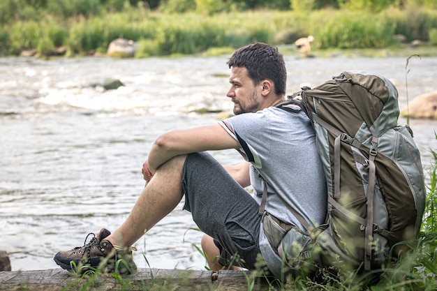 Un viajero masculino con una gran mochila de senderismo descansa cerca del río.