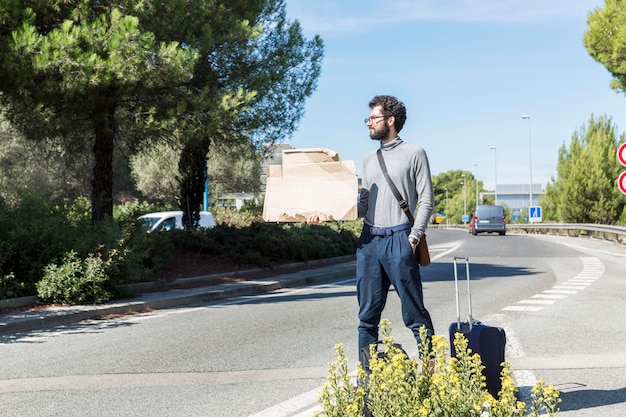 Un viajero masculino con una maleta y un cartel de cartón vacío se encuentra en la carretera Un tipo con gafas y barba Autostop gratis Espacio para texto