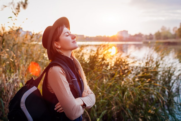 Foto viajero con mochila relajante por otoño río al atardecer. mujer joven respirando profundamente sintiéndose feliz y libre