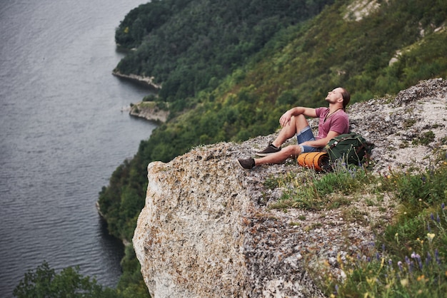 Viajero con mochila sentado en la cima de la montaña disfrutando de la vista sobre la superficie del agua. concepto de libertad y estilo de vida activo