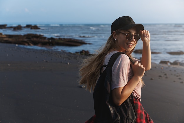 Viajero mochilero feliz, vuelve a sonreír a la cámara en una playa de arena negra.