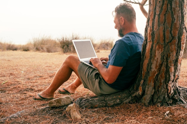 Un viajero moderno, trabajador libre, estilo de vida independiente, trabajando en una computadora portátil, sentado en el suelo contra un árbol y disfrutando de la naturaleza, conexión externa a la computadora y tecnología de trabajo para pequeñas empresas en línea