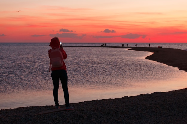 Viajero de la mujer que usa el teléfono inteligente y que toma la foto de la puesta del sol colorida del mar.