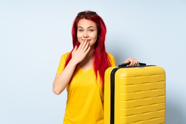 Viajero mujer sosteniendo una maleta aislada en la pared azul feliz y sonriente cubriendo la boca con la mano