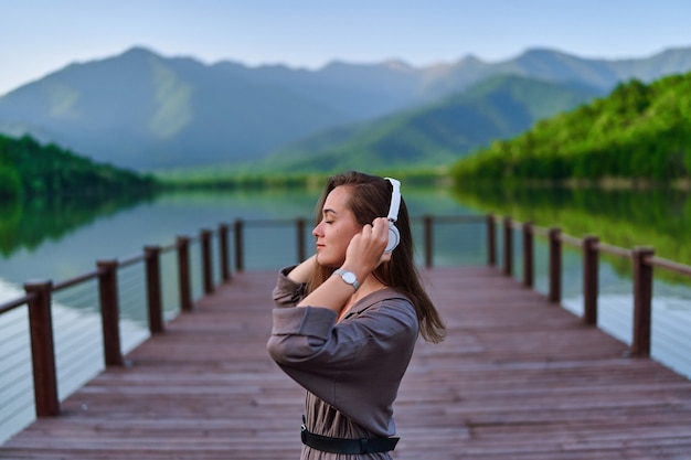 Viajero de niña escuchando música tranquila en auriculares inalámbricos de pie solo en el muelle con vista al lago y a las montañas y disfrutando de un ambiente sereno, tranquilo y pacífico