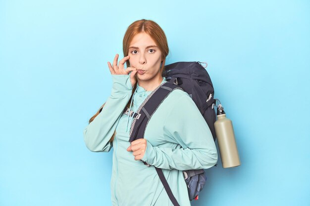 Viajero pelirrojo con mochila y colchoneta en el estudio con los dedos en los labios guardando un secreto