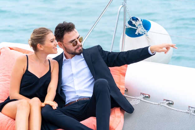 Viajero relajante pareja de lujo en bonito vestido y suite sentarse en puf en parte del yate de crucero con fondo de mar y cielo blanco. Concepto de viaje de negocios.