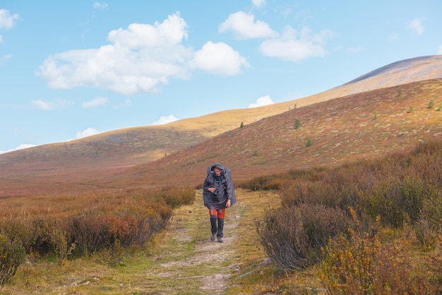 Viajero solo con impermeable y mochila grande camina por senderos entre vegetaciones otoñales que se desvanecen en la meseta de alta montaña Mochilero con cámara fotográfica en trekking de montaña en tiempo lluvioso