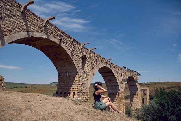 Viajero con sombrero negro se sienta junto al antiguo puente de piedra destruido en el verano