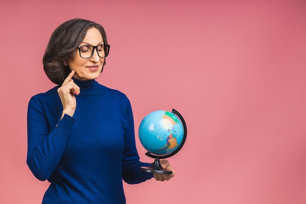 Viajero turista pensativo mujer mayor anciana madura sosteniendo en las manos Globo terráqueo aislado en el retrato de estudio de fondo rosa. Copie el espacio para el texto.