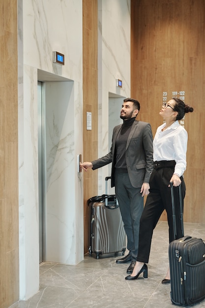 Viajeros de negocios jóvenes con equipaje de pie junto a una de las puertas del ascensor en el hotel y mirando el panel de cuenta atrás arriba