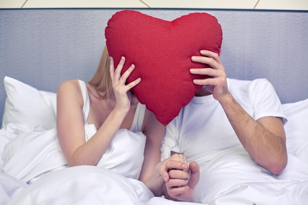 Foto viajes, amor, día de san valentín, vacaciones y felicidad concepto - pareja feliz en la cama escondiendo caras detrás de almohada en forma de corazón roja en el hotel o en casa