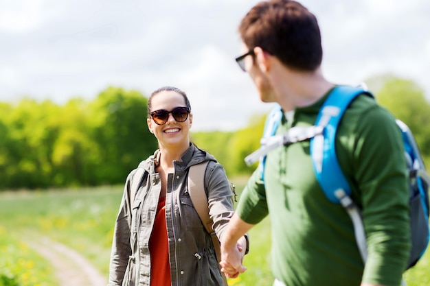 viajes, caminatas, mochileros, turismo y concepto de personas - pareja feliz con mochilas tomándose de la mano y caminando por la carretera rural al aire libre