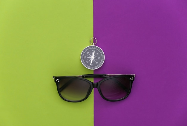 Viajes minimalistas, aventura plana. Brújula y gafas de sol sobre fondo verde púrpura. Vista superior