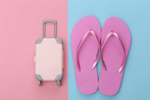 Viajes o resort de playa en plano. Mini maleta de viaje de plástico y chanclas sobre fondo rosa pastel azul. Estilo minimalista. Vista superior