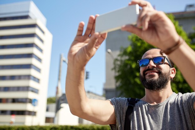 viajes, turismo, comunicación, tecnología y concepto de personas - hombre sonriente con mochila tomando video o selfie por teléfono inteligente en la calle de la ciudad de verano