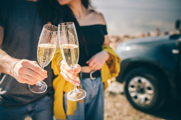 Viajes, turismo: el hombre y la mujer beben champán cerca del agua en una mesa portátil plegable. Picnic cerca del agua. Pareja de aventuras. Concepto de viaje en coche.