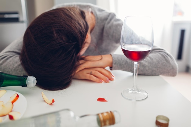 Vício em álcool feminino Jovem dormindo na mesa da cozinha cercada de garrafas de vinho