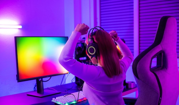 Victoria ganadora jugadora asiática jugando videojuegos en línea emocionada en computadora de escritorio PC coloridas luces LED de neón mujer joven con auriculares para juegos usando computadora ella feliz exitoso concepto de deporte electrónico