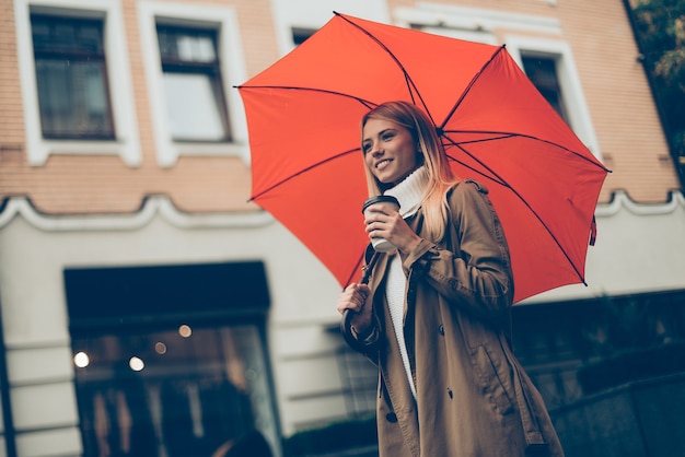 Vida en la ciudad. Vista de ángulo bajo de la atractiva joven mujer sonriente que lleva paraguas y una taza de café mientras camina por la calle