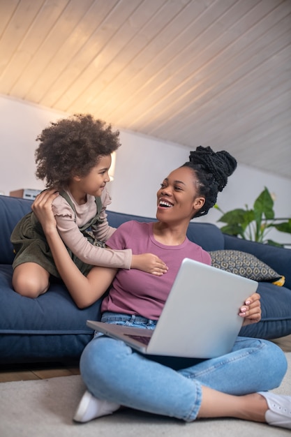Vida cotidiana. Alegre mãe afro-americana com laptop sentado no chão e uma menina tocando, exigindo atenção em casa