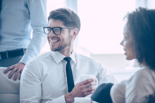 Vida de empresario. Primer plano de joven guapo alegre sosteniendo la taza de café y mirando a otro lado con una sonrisa mientras está sentado en la oficina con sus compañeros de trabajo