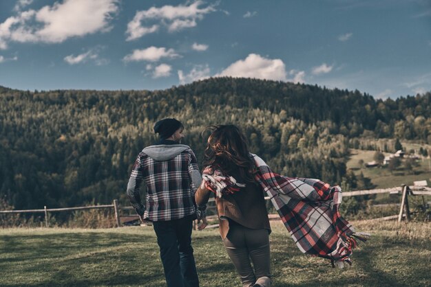 ¡La vida es asombrosa! Vista trasera de la joven pareja juguetona corriendo mientras pasa tiempo sin preocupaciones en el valle en las montañas