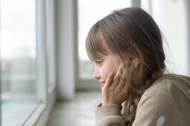 Vida sin esperanza. Cerca de una pobre niña deprimida parada cerca de la ventana y mirando a un lado mientras se siente miserable