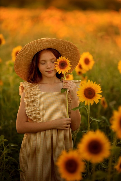 Vida feliz no retrato de verão de uma linda garota ruiva com um chapéu em um campo de girassóis com flores nas mãos nos raios do sol poente