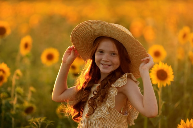 Vida feliz en el retrato de verano de una hermosa chica pelirroja con un sombrero en un campo de girasoles con flores en las manos en los rayos del sol poniente