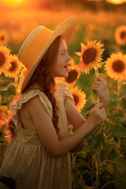 Vida feliz en el retrato de verano de una hermosa chica pelirroja con un sombrero en un campo de girasoles con flores en las manos en los rayos del sol poniente