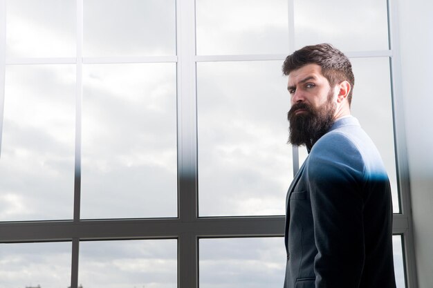 Vida moderna. hombre de negocios de pie en la ventana con paisaje urbano. Hombre barbudo brutal. Hipster maduro con barba. Hombre en la oficina de negocios moderna. Hombre de negocios en traje formal. Vida moderna del empresario.