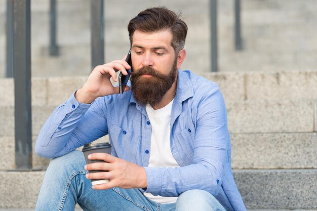 Vida móvel Hipster bebe café falando no celular ao ar livre Comunicação móvel Copo quente Café para viagem Nova tecnologia Dispositivo móvel 4G 3G Estilo de vida moderno