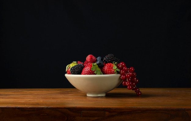 Vida muerta de frutas de bayas en cuencos en una mesa de madera contra un fondo negro
