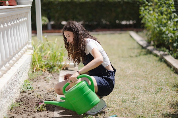 Vida suburbana Jovem mulher com cabelo encaracolado trabalhando no quintal regando flores no jardim