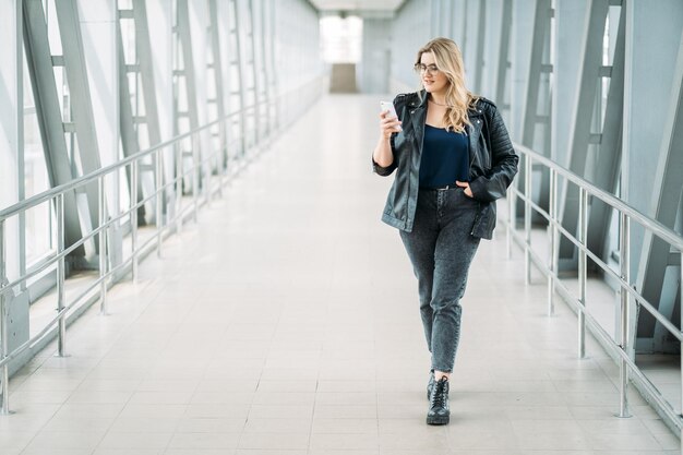 Vida urbana Cuerpo positivo Comunicación móvil Gente de gadgets Mujer obesa de talla grande con sobrepeso elegante y segura que usa mensajes de texto telefónicos caminando sola en un puente moderno