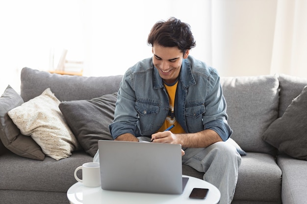 Video chamada. Jovem homem milenar caucasiano se comunica em redes sociais ou trabalha on-line usando um laptop, sentado no sofá de casa