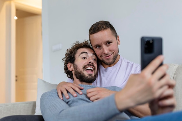 Vídeo de casal gay bonito chamando seus amigos na sala de estar em casa Dois amantes do sexo masculino sorrindo alegremente enquanto cumprimentam seus amigos em um smartphone Jovem casal gay sentados juntos