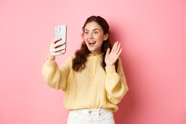 Vídeo de menina alegre conversando no smartphone acenando com a mão para a câmera do telefone e sorrindo feliz dizendo olá em pé contra a parede rosa