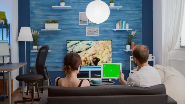 Vídeo estático de casal olhando para tablet digital com tela verde assistindo conteúdo de vídeo online na frente da tv. Homem e namorada segurando o dispositivo touchscreen com chroma key curtindo o vlog do influenciador.