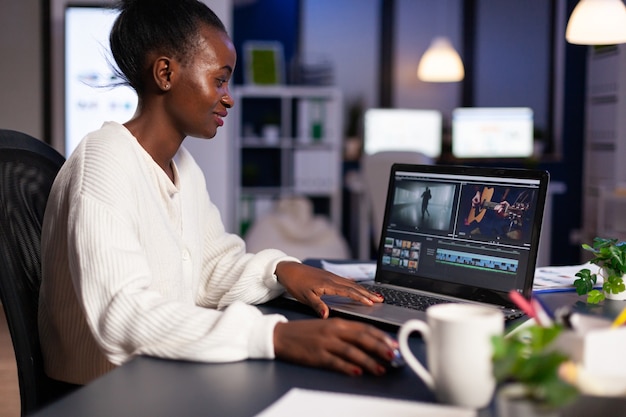 Vídeo negro encantado editando filme em um laptop profissional sentado na mesa de um escritório à meia-noite