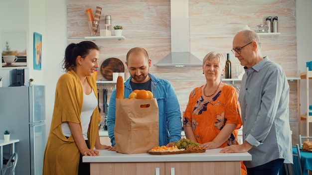 Video retrato de familia extensa feliz sonriendo a la cámara, sentado en la cocina. Personas en el comedor alrededor de la bolsa de papel con comestibles mirando la cámara web