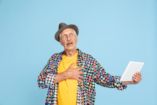 Videochat usando tableta, sorprendido. Retrato de hombre senior hipster con elegante sombrero en azul. Tecnología y concepto de estilo de vida de ancianos alegre