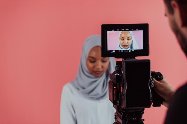 Videógrafo en un estudio digital grabando video en una cámara profesional disparando a una mujer musulmana usando un pañuelo hiyab de fondo rosa de plástico. foto de alta calidad