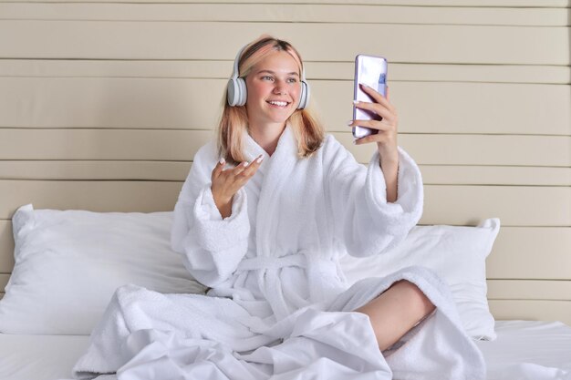 Videollamada de comunicación en línea adolescente en auriculares con teléfono inteligente riendo y hablando Mujer sentada en la cama en bata de baño blanca descansando charlando con amigos usando comunicación por video