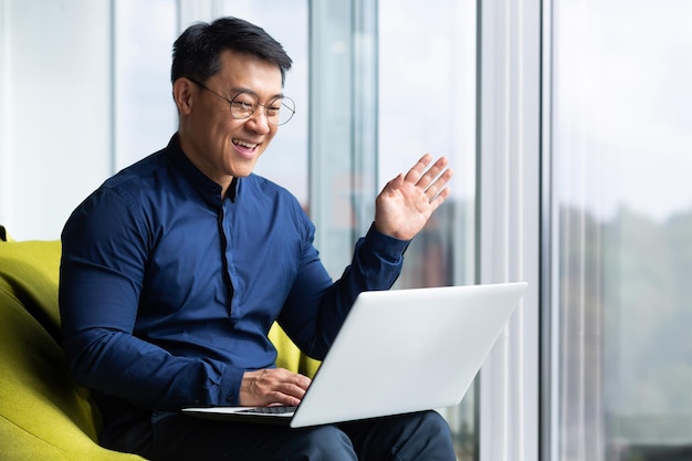 Videollamada exitoso hombre de negocios asiático cerca de la ventana en la oficina hablando remotamente en línea con