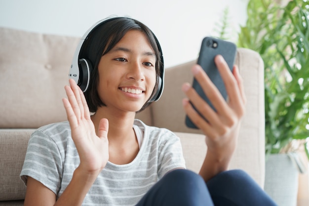 Videollamada de niña asiática con un auricular en el teléfono inteligente y saludando mientras está sentado en la sala de estar