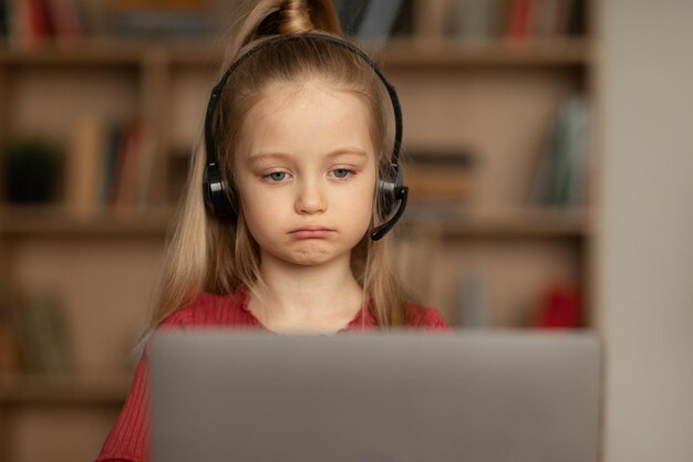 Videollamadas de colegiala poco infeliz a través de una computadora portátil aprendiendo en línea en el interior