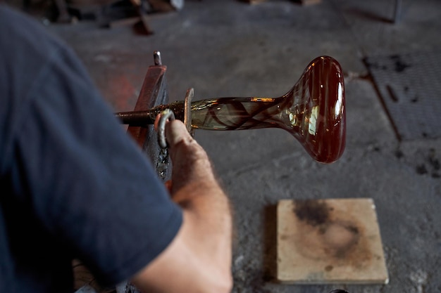Vidriero masculino irreconocible en proceso de hacer un jarrón de narguile usando cerbatana y pinzas mientras trabajaba en una fábrica de soplado de vidrio