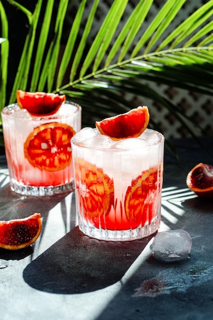 Vidrio con agua mineral fría con naranjas sanguinas y cubitos de hielo sobre fondo de hormigón Sombra de hoja de palma tropical Fondo de luz solar y sombra